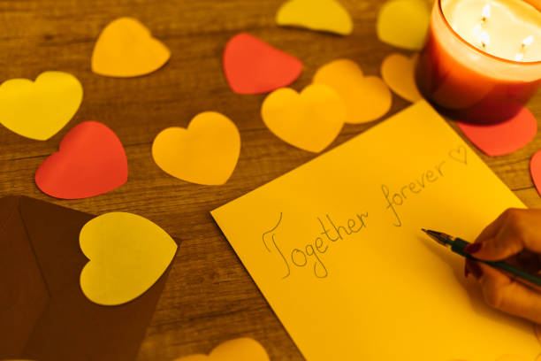 A hand is composing a heartfelt message on paper surrounded by vibrant paper hearts and an atmospheric candle for a Valentine's Day. The scene evokes feelings of love, romance, and creativity through warm tones and a personal touch