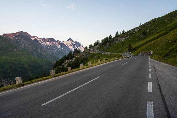 Asphalt road winds among alpine meadows, cliffs, leading to majestic Grossglockner mountain. Snow capped peaks on horizon. For themes of travel, nature, outdoor activities.