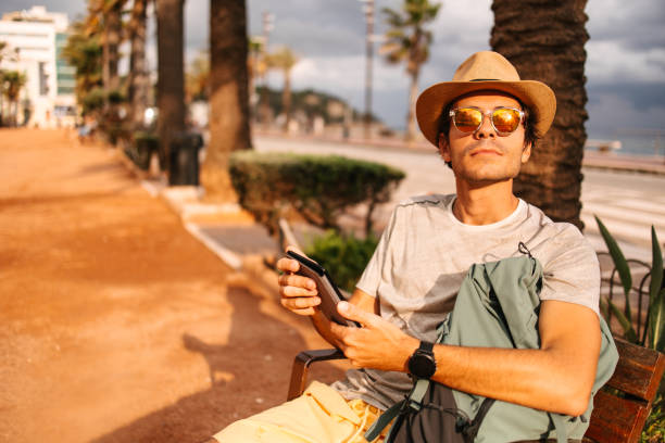 A young male tourist with a hat, backpack, and sunglasses, sitting on a bench in Spain, reading on an electronic reader. Clouds and sun