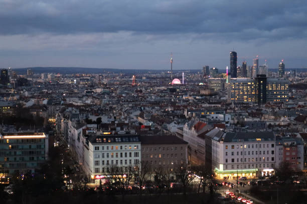 A mesmerizing aerial view of Vienna at twilight, where the city's historic architecture and modern skyline blend harmoniously under the fading evening light. The soft glow of streetlights illuminates the elegant streets, while the Danube River reflects the warm hues of dusk. This breathtaking cityscape captures the timeless charm of Austria’s capital from above