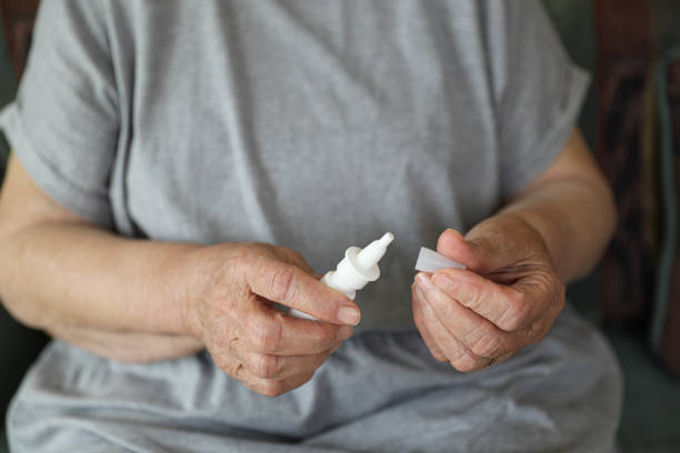 The image shows an elderly woman sitting comfortably in a cozy, well-lit room, holding a nasal spray in her hand. She appears to be in the middle of using it or preparing to administer a dose. Her facial expression conveys relief and focus as she carefully follows the instructions.
