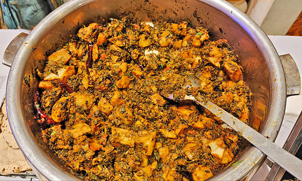 A large stainless steel pot filled with spiced potato and fenugreek curry, featuring golden-brown potato cubes mixed with cooked fenugreek leaves, whole dried red chilies, and aromatic Indian spices, with a serving spoon resting inside.