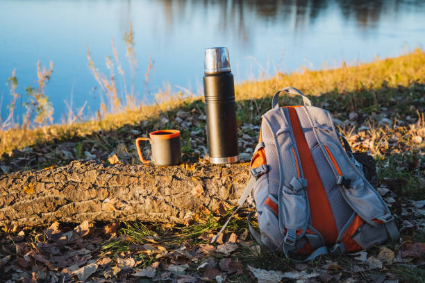 A sturdy backpack, an insulated thermos, and a cozy mug are resting on the ground close to a serene lake, surrounded by beautiful natural scenery and lush grass that enhances the landscape