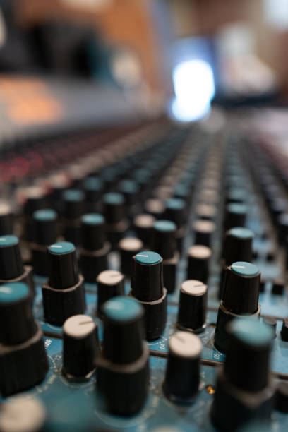 A detailed view showcases a sound mixing console filled with black and teal knobs arranged neatly in rows within an active recording studio. Soft lighting creates a focused atmosphere.