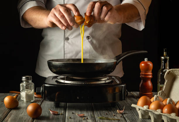 istock A chef, dressed in a white uniform, skillfully cracks eggs into a black frying pan. The warm light highlights the fresh ingredients and kitchen utensils arranged on a wooden countertop 2198187765