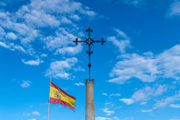 A decorative metal cross stands on a stone pillar with a bright blue sky and a Spanish flag waving in the wind beside it, highlighting details.