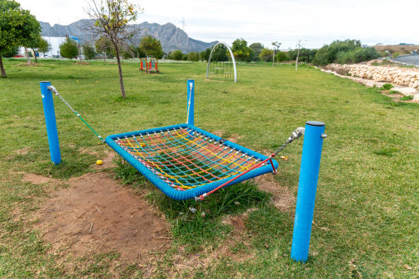 A colorful rope hammock with a blue frame is secured between posts on a green playground. Grass and small trees surround the play area under a cloudy sky.