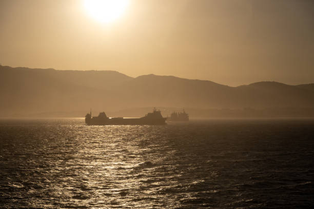 Two cargo ships near a hilly coastline during sunset; the scenery is bathed in golden and orange tones.