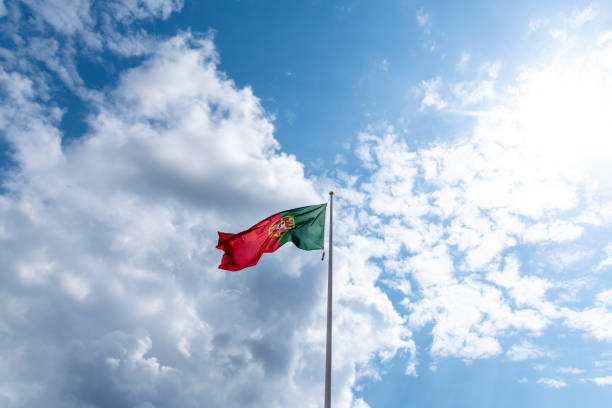 The flag of Portugal, waving in the breeze on a sunny day. The flag design has red and green color blocks, with an armillary sphere (representing the country's Age of Discovery), shields (representing victories), and castles (representing historic independence) decorating a central circular coat of arms area. Officially adopted in 1911, the green symbolizes hope, while the red stands for the blood of patriots who defended the country. The national flag of the Portuguese Republic is shown attached to a flagpole against a clear blue sky, with copy space.