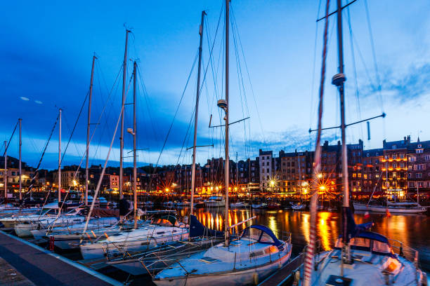 Honfleur, France. July 27th, 2019: A beautiful evening view of the marina in Honfleur, with sailboats gently swaying in the water. The warm glow of the town’s lights reflects on the calm surface, contrasting with the deep blue of the twilight sky.