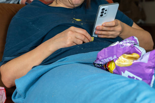 A person lounging on a couch, eating chips, and using a smartphone, with snack packaging scattered around. Depression.