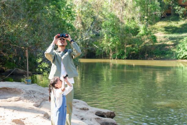 istock Asian father and daughter standing by a river, both holding binoculars observing nature. Outdoor learning, wildlife appreciation, and childhood discovery in a green and peaceful setting. 2198211257