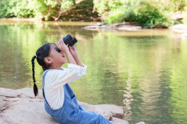 istock Asian girl sitting by a river looking up through binoculars, engaged in nature exploration. Childhood curiosity, adventure, and outdoor learning experience in a green and peaceful environment. 2198211275