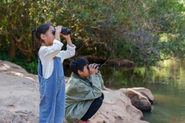 istock Asian brother and sister exploring nature by a river using binoculars. Childhood curiosity and outdoor learning in a peaceful green environment, encouraging adventure and discovery. 2198211281