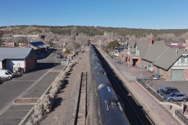 Aerial view of freight train traveling through mountainous terrain in Storey County, Nevada. Train tracks curve alongside barren hills with small rural settlement visible in foreground. Scattered trees provide shade for residential buildings in otherwise dry landscape. Dirt roads connect properties across desert scrubland. Truckee River valley visible with mountains rising in background under partly cloudy sky. High angle view captures transportation infrastructure cutting through rugged Nevada wilderness.