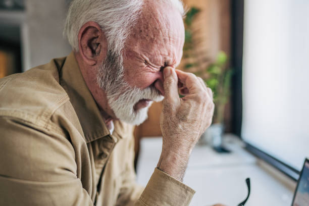 istock An senior man stands in the kitchen, using his laptop. His expression is strained as he furrows his brow and grips his head with one hand, showing signs of discomfort. 2198253916