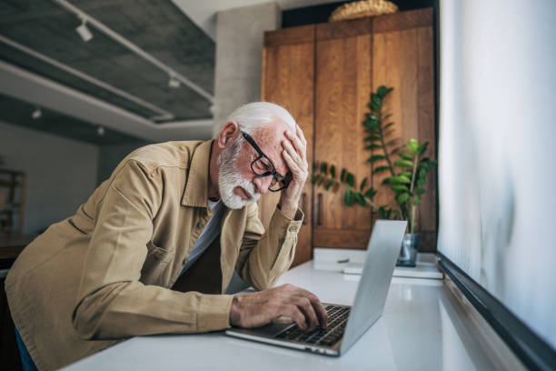 An senior man stands in the kitchen, using his laptop. His expression is strained as he furrows his brow and grips his head with one hand, showing signs of discomfort.