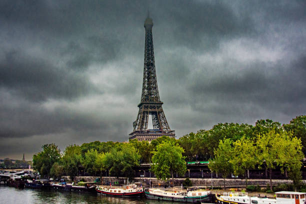 The Eiffel Tower rises behind lush green trees and the Seine River, with colorful riverboats docked below, under an overcast sky in Paris, France.