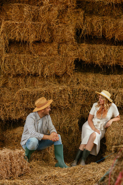 Two young adults sitting and chatting in front of stacked hay bales on a farm. They are wearing casual farming attire and appear to be enjoying a break