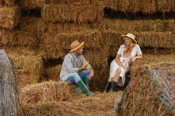 Two adults sitting on hay bales in a barn, engaging in a friendly conversation. Both wearing hats, suggesting a rural or farming lifestyle
