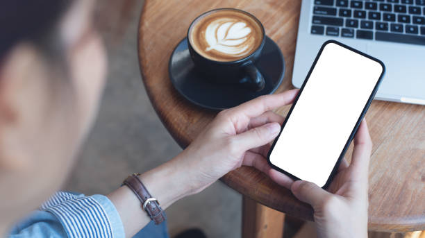 istock Cell phone white screen mockup, Woman hand holding mobile phone with blank screen while working at coffee shop. Business woman using smartphone, mockup for social media advertisement 2198297077