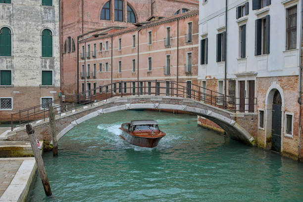 A lone boat in Venice glides under a charming stone arched bridge. The turquoise canal flows between historic buildings with green shutters, reflecting centuries of Venetian charm.