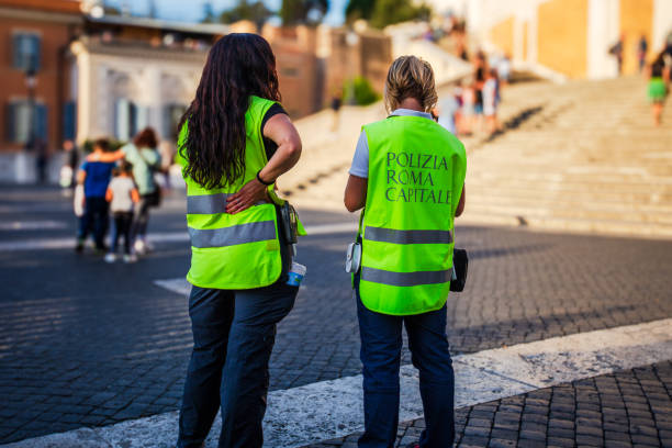 Rome, Italy. October 12th, 2019: Two officers of the Polizia Roma Capitale wearing bright yellow safety vests, standing in conversation on a cobblestone street near a busy tourist area. The scene captures the blend of local law enforcement and vibrant city life in the heart of Rome.