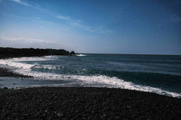 A striking volcanic pebble beach in Lanzarote, Canary Islands, where dark, smooth stones contrast with the deep blue Atlantic Ocean. The rugged coastline, shaped by volcanic activity, meets powerful waves crashing against jagged rocks. A dramatic and untamed seascape, showcasing the raw beauty of nature.