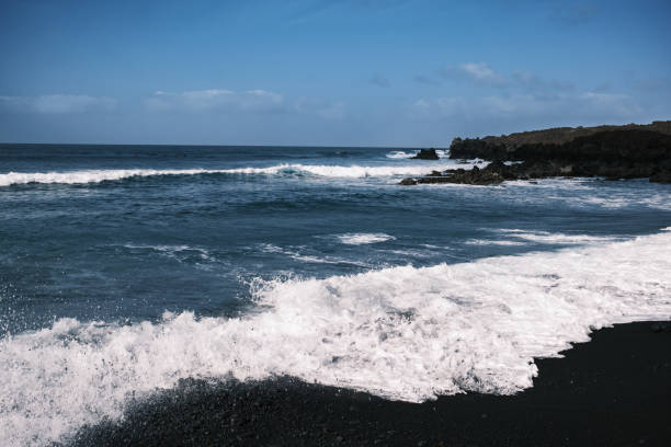A striking volcanic pebble beach in Lanzarote, Canary Islands, where dark, smooth stones contrast with the deep blue Atlantic Ocean. The rugged coastline, shaped by volcanic activity, meets powerful waves crashing against jagged rocks. A dramatic and untamed seascape, showcasing the raw beauty of nature.