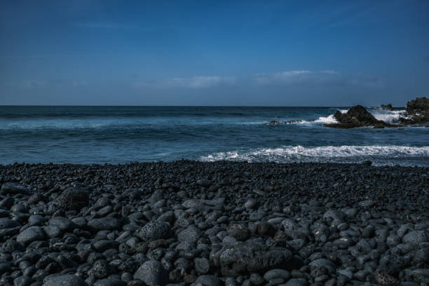 A striking volcanic pebble beach in Lanzarote, Canary Islands, where dark, smooth stones contrast with the deep blue Atlantic Ocean. The rugged coastline, shaped by volcanic activity, meets powerful waves crashing against jagged rocks. A dramatic and untamed seascape, showcasing the raw beauty of nature.