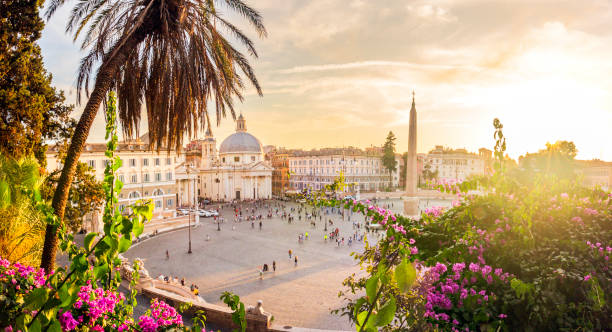 Rome, Italy. October 13th, 2019: A breathtaking view of Piazza del Popolo during a golden sunset, framed by vibrant flowers and lush greenery. The square's iconic obelisk and the twin churches, Santa Maria dei Miracoli and Santa Maria in Montesanto, shine in the warm evening light, creating a serene yet lively atmosphere.