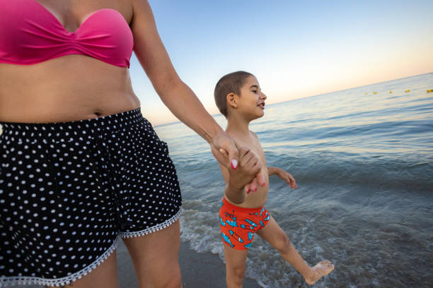 A loving mother and her son with a mental disability walk hand in hand along the shoreline, enjoying a peaceful vacation by the sea.