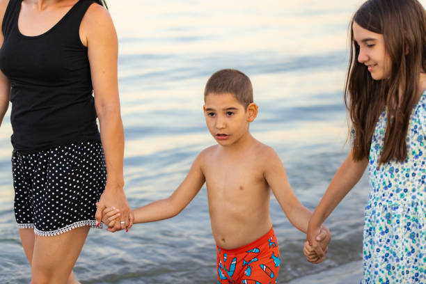 Holding hands and embracing the moment, a mother, daughter, and little boy with a mental disability enjoy a serene beachside stroll together.