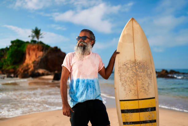 istock Active senior man with surfboard on sandy beach. Older surfer enjoys sunny day by ocean. Retirement goals, fitness, freedom lifestyle near waves. Happy golden years, surf life, beach adventure. 2198339421