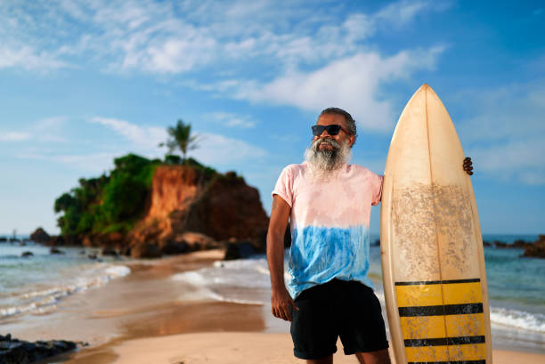 Elderly man with surfboard on sunny beach. Active senior enjoying ocean view, healthy retirement lifestyle adventure. Surfing as leisure in golden years. Celebrating vitality, new experiences.