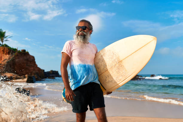 istock Smiling senior man with beard holds surfboard on sunny beach. Active older surfer in sunglasses enjoys ocean waves. Travel destinations for surfing. Healthy lifestyle. 2198341581