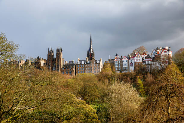 Edinburgh skyline featuring historic architecture and gothic spires, framed by lush green trees under a dramatic cloudy sky