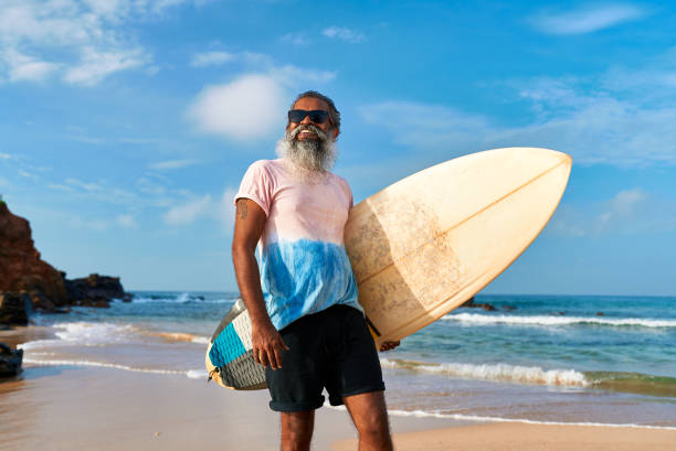istock Senior ethnic man with surfboard on sandy beach. Active lifestyle, ready to surf ocean waves. Older surfer enjoys sunny travel destination. Inspires adventure tourism, healthy senior activities. 2198343421
