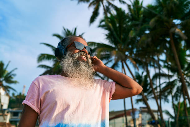 istock Gray-bearded elderly man dances joyfully on tropical beach with wireless headphones. Stylish senior enjoys music under palm trees, wearing sunglasses. Vibrant summer vibe. Beach party poster. 2198344724
