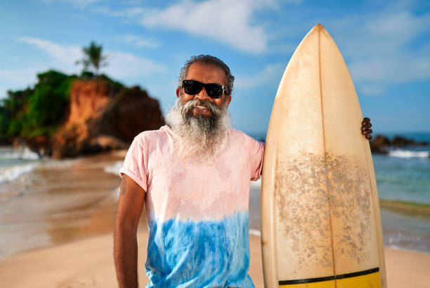 istock Senior bearded man with surfboard on sunny beach. Elderly surfer enjoys active lifestyle by ocean. Older male in sunglasses embraces sea adventures and fitness. Travel, leisure vibes. 2198346134