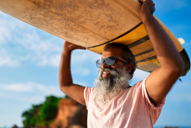 istock Active senior ethnic man smiles holding surfboard on sunny beach. Retired surfer enjoys ocean sport. Elderly embraces adventure, free lifestyle in nature. Healthy aging and outdoor fun. 2198347251