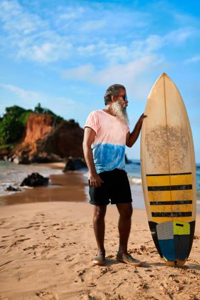 istock Senior man with surfboard stands on sandy beach. Elderly surfer poses under blue sky by rocky coast. Active retiree enjoys outdoor sea adventure. Tropical surf lifestyle travel concept. 2198351879