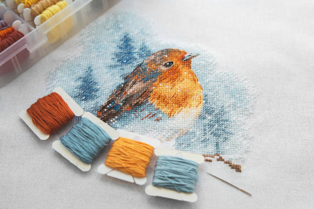 top view of the cross stitch process multicolored bird, white canvas, floss threads on bobbins, needle, needlework and cross stitch concept 1
