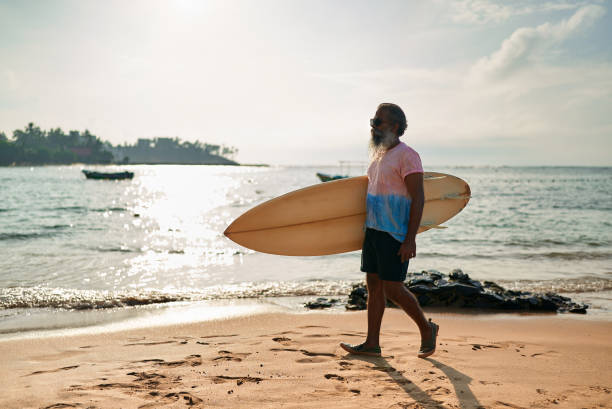 istock Active senior ethnic man walks on sunny beach with surfboard. Elderly surfer enjoys ocean waves in vibrant lifestyle. Retirement adventure with surf culture. Promoting healthy aging in coastal scene. 2198357436