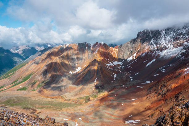 istock Top view from rocks to rounded multicolor valley with curved iron river and big sharp rocky ridge of red color. Colorful large mountain range in freshly fallen snow. Vivid high mountains in low clouds 2198357880