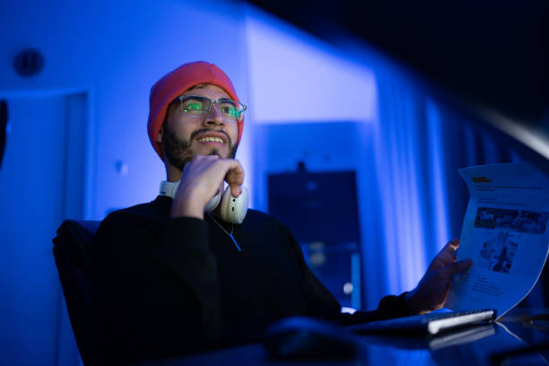 istock Young man with glasses wearing a beanie engages with documents while seated at a desk in a blue-lit room 2198358741