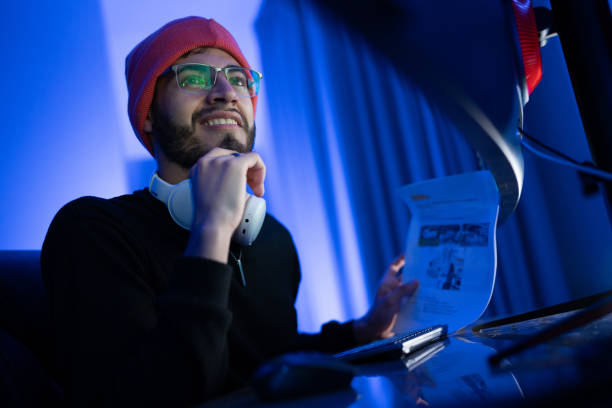 Young man with glasses and headphones smiles while reading a document in a dimly lit room with blue ambiance