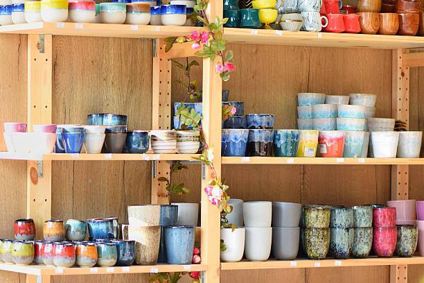 Various handmade ceramic cups and small pots arranged on wooden shelves, surrounded by decorative artificial flowers. A vibrant mix of colors, textures, and glaze patterns, ideal for showcasing artisan crafts or home decor.