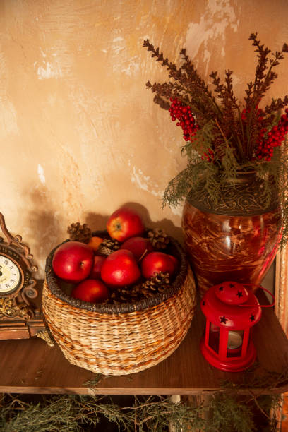 A wicker basket full of red apples. Nearby is a decorative red lantern and a vase with pine branches.