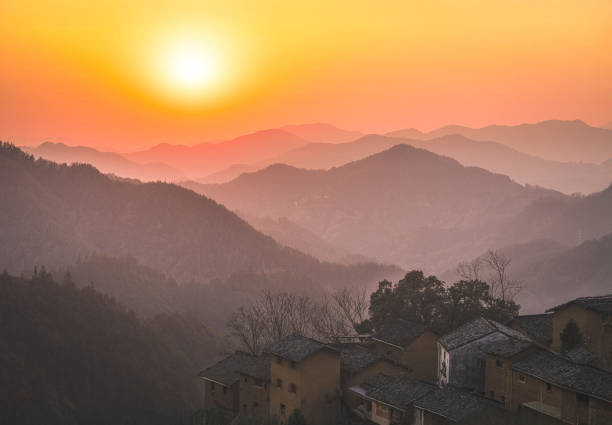 A tranquil sunset over a mountainous village, where layers of rolling hills fade into the warm hues of the evening sky. The sun, glowing like a radiant orb, bathes the scene in a golden-orange light, creating a mesmerizing gradient that transitions from deep amber near the horizon to soft pastel tones above.\n\nNestled in the foreground, a cluster of traditional hillside houses with weathered clay walls and dark-tiled roofs adds a sense of history and cultural depth.
